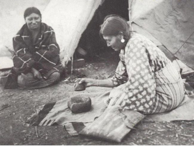 Two Native American women sitting outside a tent, one grinding mesquite beans with a stone on a mat