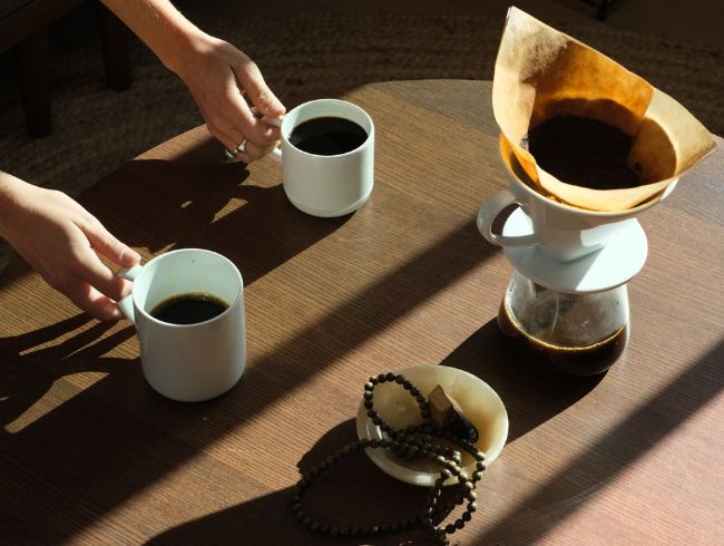 Pour-over mesquite coffee setup on a wooden table with two mugs, prayer beads, and a bowl in warm sunlight.