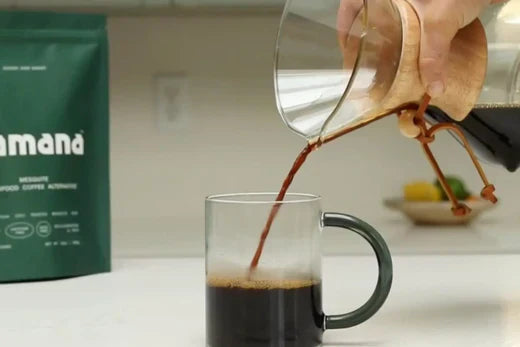 Hand pouring brewed mesquite coffee from a Chemex into a glass mug beside Kamana mesquite coffee packaging