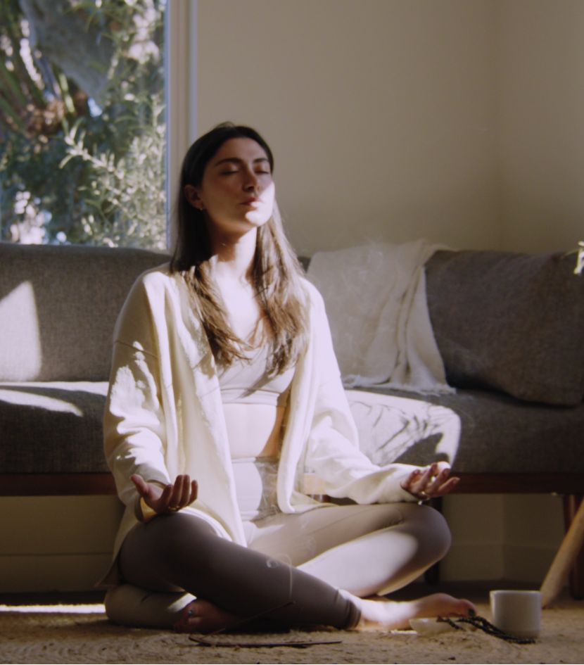 Woman meditating in a sunlit room, sitting cross-legged on the floor in a peaceful & relaxed posture